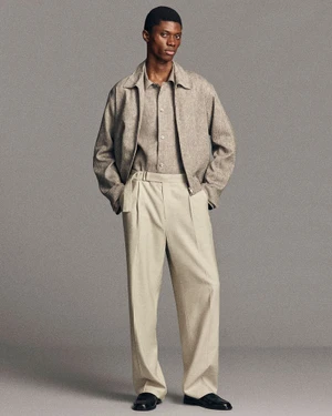 Black man wearing a beige jacket, matching shirt, and light trousers standing against a solid grey studio background.