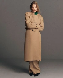 Model standing against a grey seamless backdrop wearing a long camel coat and matching trousers with a green scarf