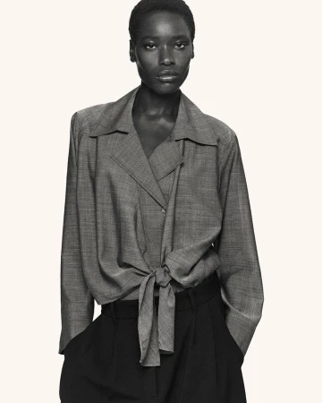Model wearing a grey long-sleeved tie-front blouse and dark trousers against an off-white background for COS Spring 2026