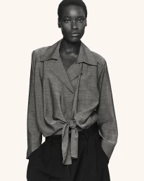 Model wearing a grey long-sleeved tie-front blouse and dark trousers against an off-white background for COS Spring 2026
