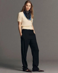 Model standing against a grey studio backdrop wearing a cream top, dark patterned neck scarf, and black trousers