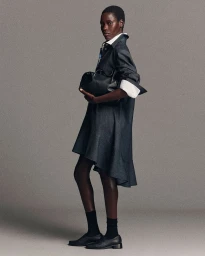 Model in dark denim dress holding black leather bag against a neutral grey studio background