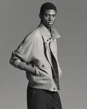 Black male model wearing a structured beige jacket and dark trousers against a grey seamless background