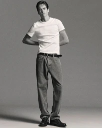 Full-length portrait of a male model wearing a white t-shirt, grey jeans, and black shoes against a grey seamless background.