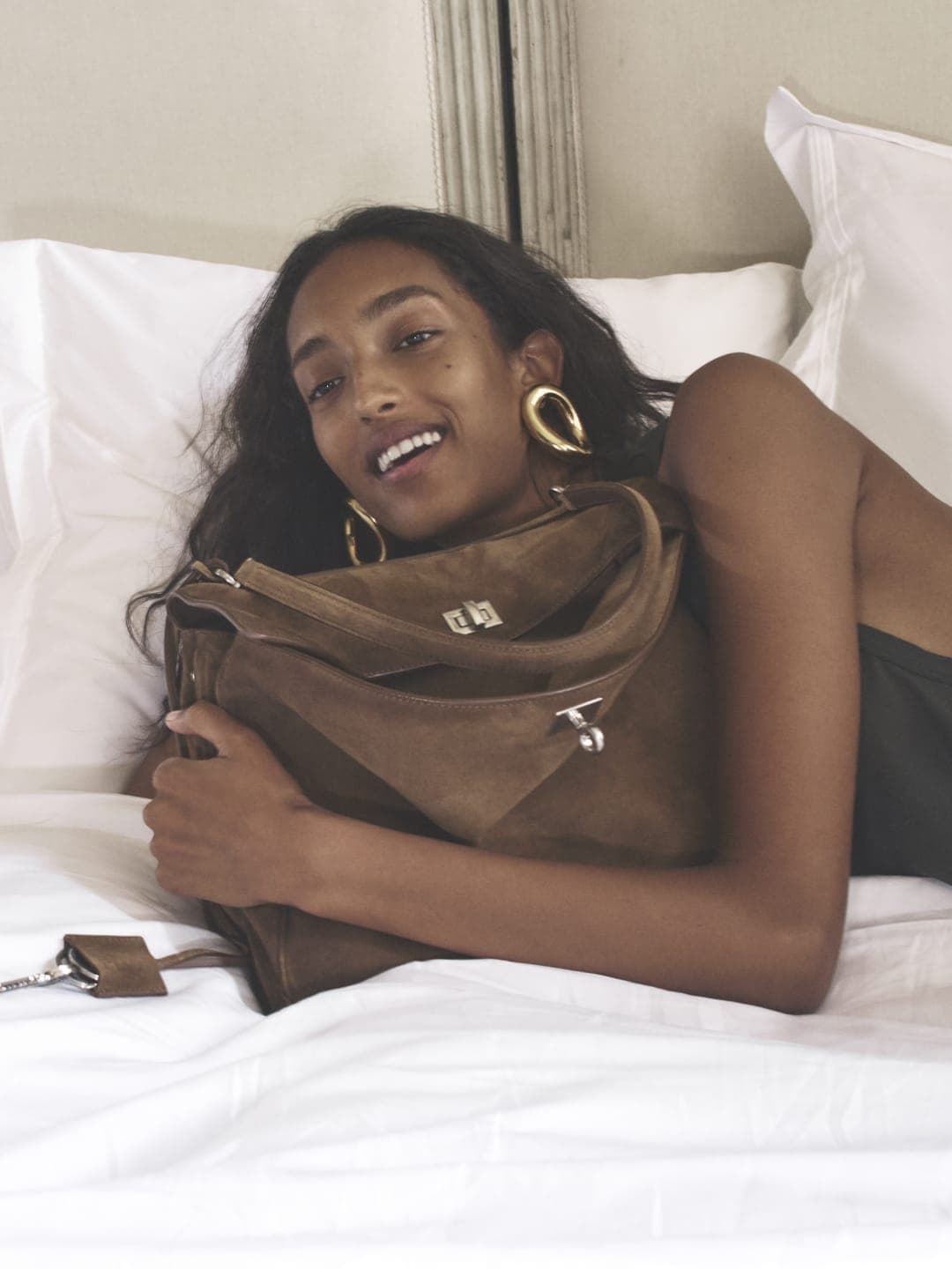 Dark-skinned woman with gold earrings and brown purse lying on a bed with white sheets in an indoor setting with natural light.