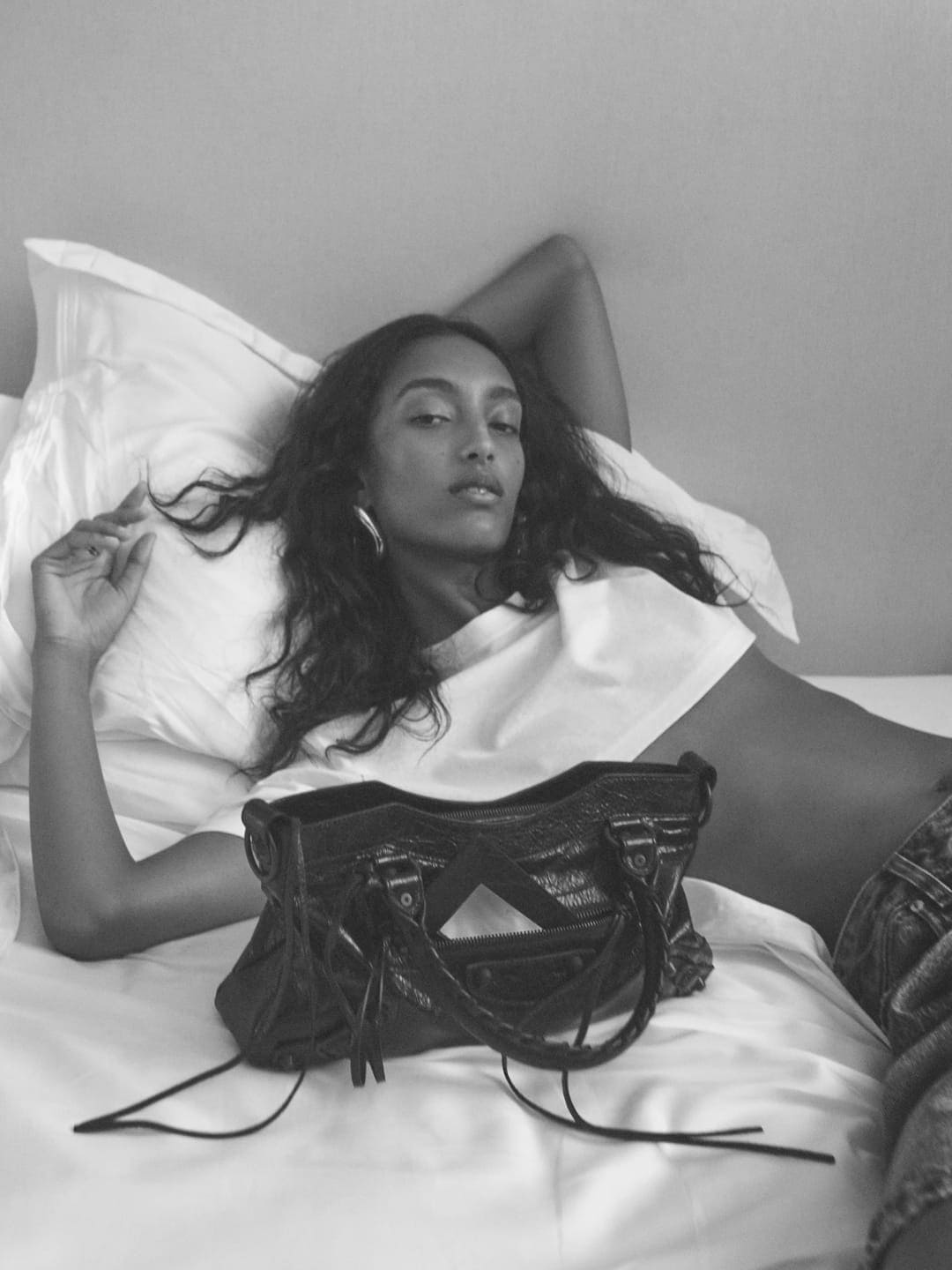 Model lying on a white bed with a black handbag in the foreground for Balenciaga Fall 2025 campaign