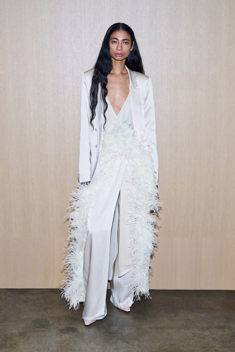 Full-length shot of a woman with long dark hair wearing a long white coat and jumpsuit with feather trim standing indoors.