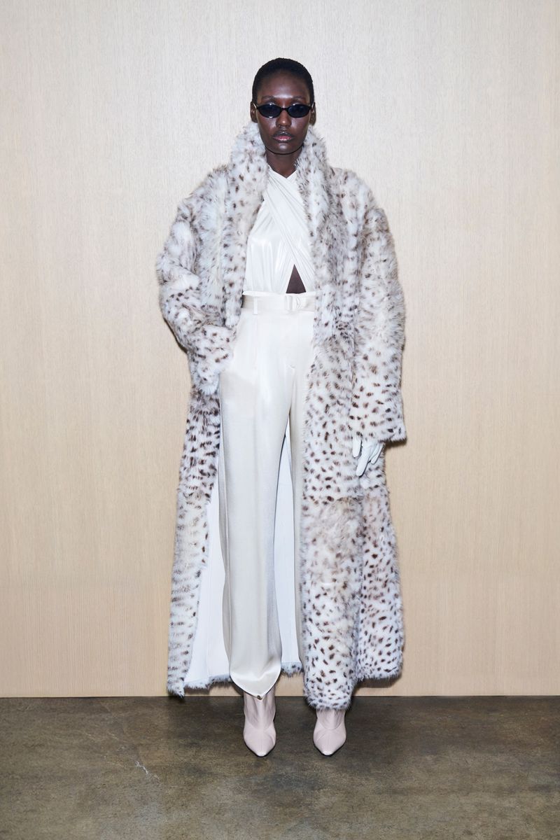 A striking fashion shot of a woman in a spotted coat and cream-colored outfit standing against a neutral backdrop, embodying modern elegance.