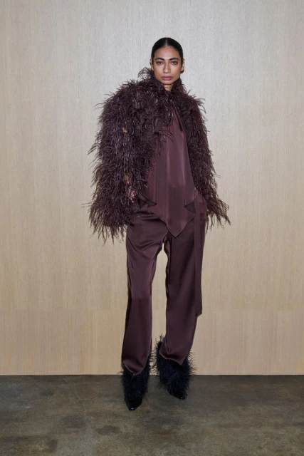 Elegant woman in a brown satin suit with a feathered jacket and feathered heels stands against a light brown wall in a studio setting.