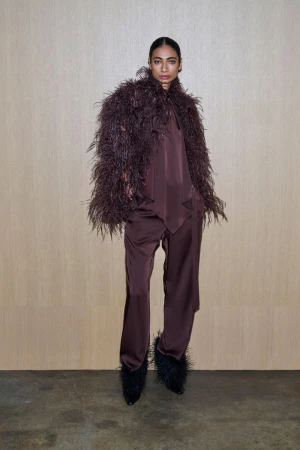 Elegant woman in a brown satin suit with a feathered jacket and feathered heels stands against a light brown wall in a studio setting.