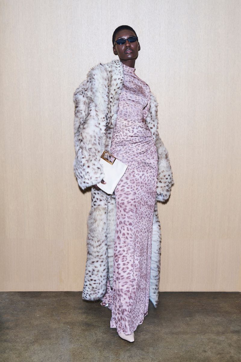 Adult model wearing a leopard print dress and white, faux-fur coat poses indoors against a beige background. She holds a white clutch.