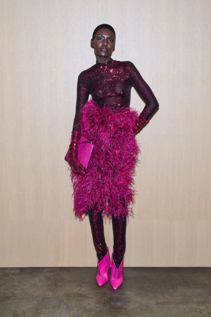 An adult female model in a sequined catsuit and pink feather skirt poses indoors against a neutral wall, holding a pink clutch and wearing pink boots.