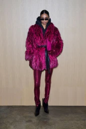 Model in magenta faux fur coat and metallic leggings wearing dark sunglasses against a wooden background