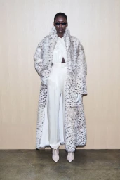 Model wearing a faux fur animal print coat and cream satin trousers against a pale wooden wall