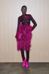 Model wearing a sequined long-sleeve top, vibrant feather skirt, matching gloves, and pink boots against a wood-paneled wall