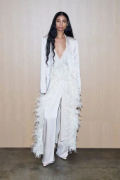 Model wearing a white satin blazer and feather-trimmed trousers against a light wood paneling background
