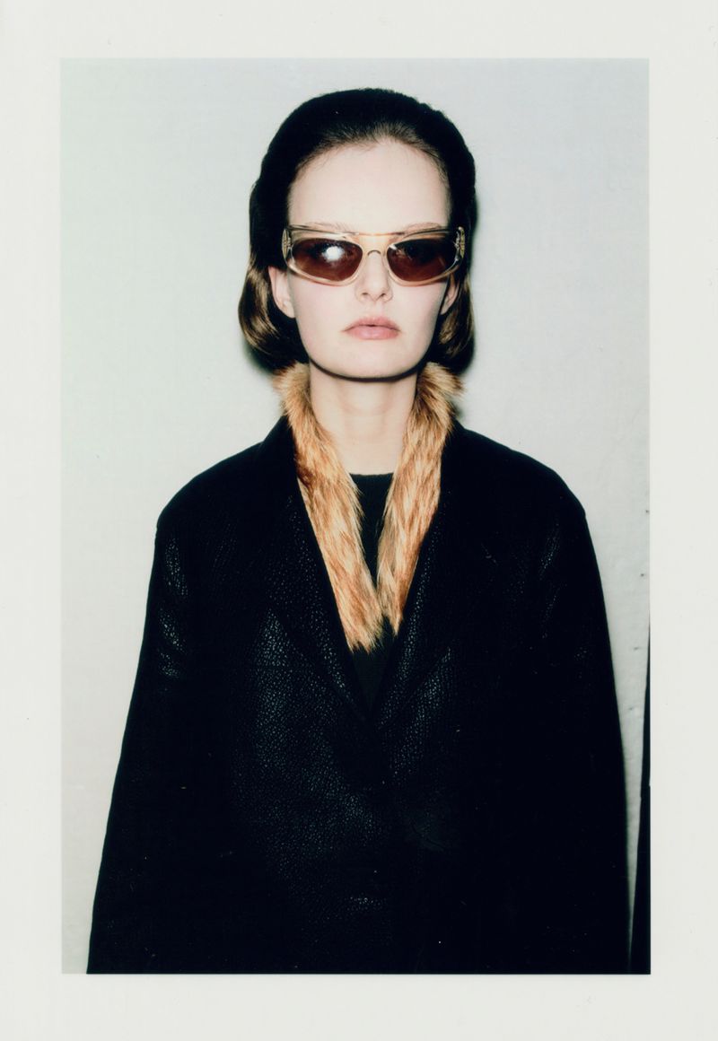 Portrait of a young woman with light skin wearing sunglasses, a dark coat with a fur collar, and a hat, against a neutral background.