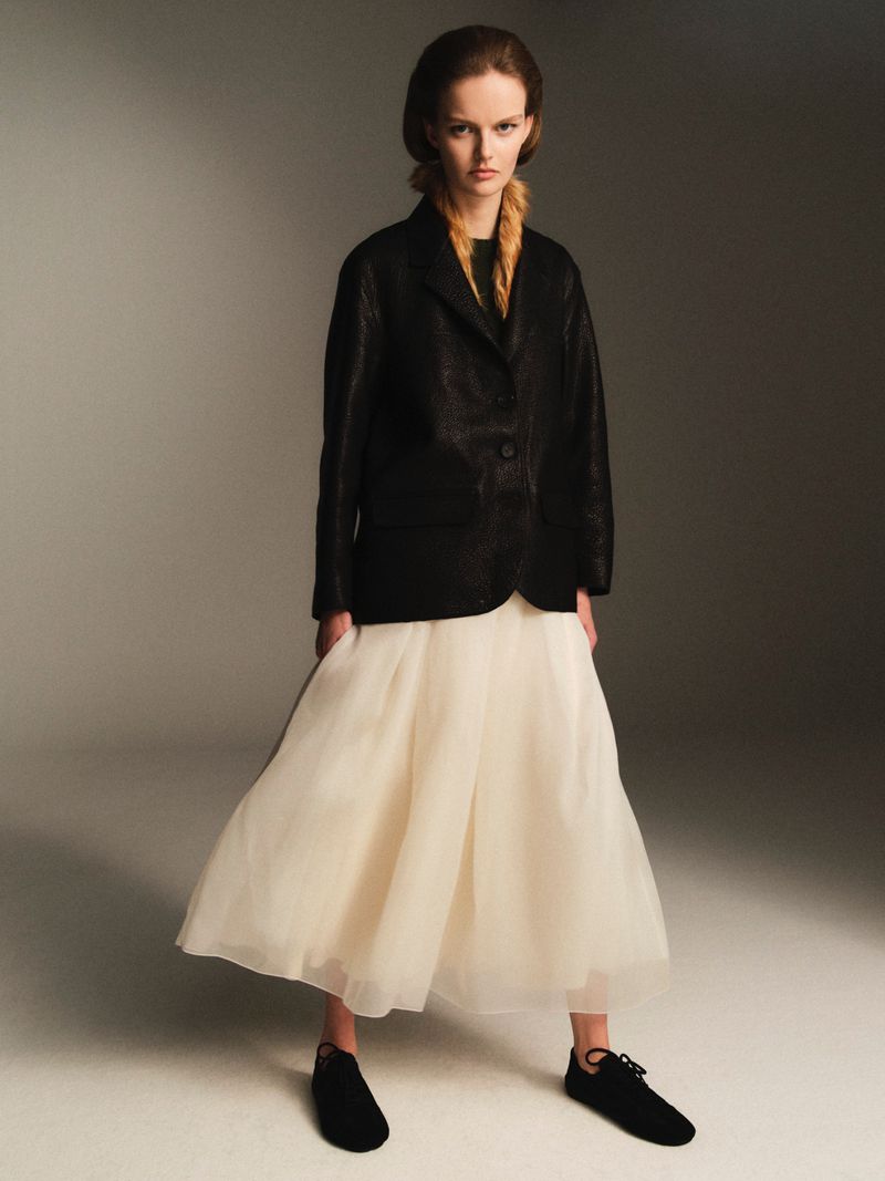 Fashion shot of a woman in a black blazer and white skirt posing in a studio, with braided hair and black flats, against a gray backdrop.