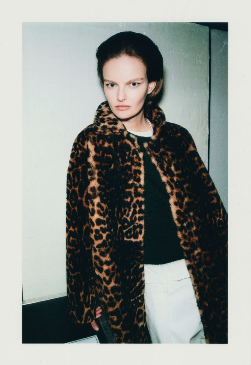 A woman poses indoors wearing a brown and black leopard print coat, a dark shirt, and white pants, looking directly at the camera.