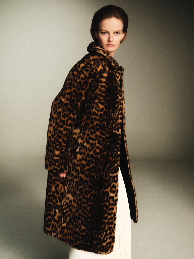 Model wearing a leopard print faux fur coat over white trousers against a neutral grey studio backdrop