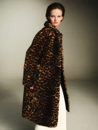 Model wearing a leopard print faux fur coat over white trousers against a neutral grey studio backdrop