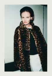 Model wearing a leopard print faux fur coat and white trousers against an indoor wall background.
