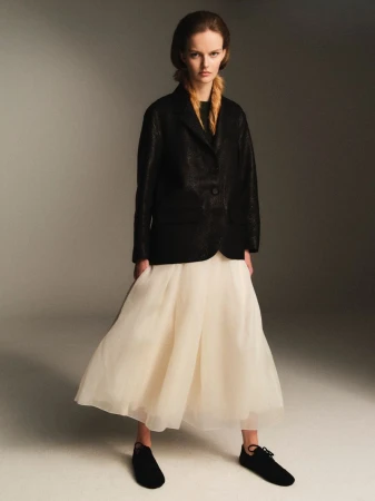 Model in a dark blazer and cream tulle skirt standing against a grey studio backdrop for Rika Studios A/W 26 lookbook.