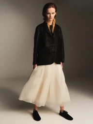 Model in a dark blazer and cream tulle skirt standing against a grey studio backdrop for Rika Studios A/W 26 lookbook.