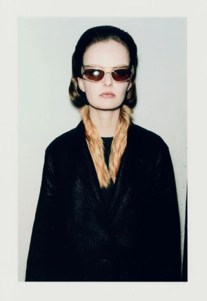 Model wearing dark sunglasses and a black coat with a faux fur collar against a plain studio background.