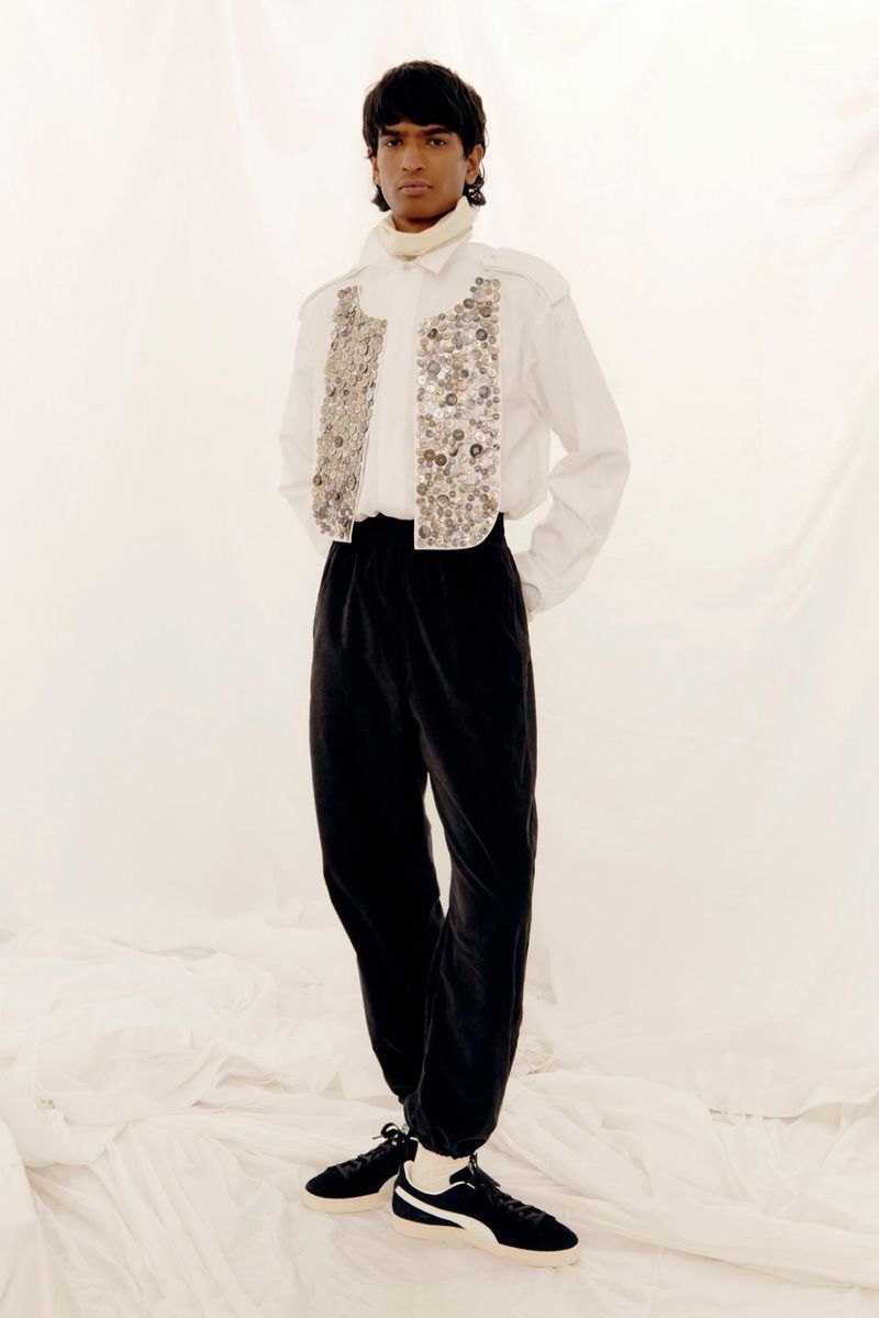 Full body studio shot of a person wearing a white button down shirt with silver embellishments and black pants against a white background.