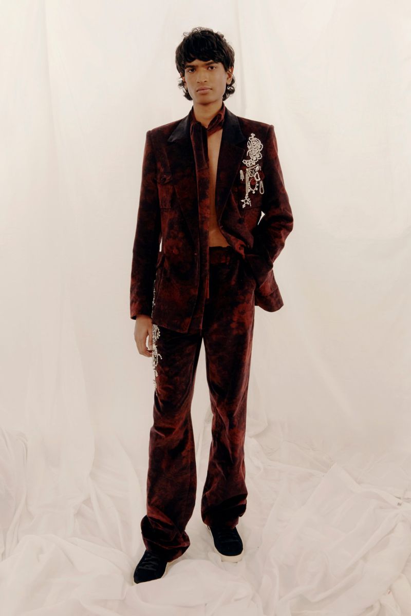 A stylish male model presents a red velvet suit with ornate silver brooches, set against a soft, neutral backdrop in a studio setting.