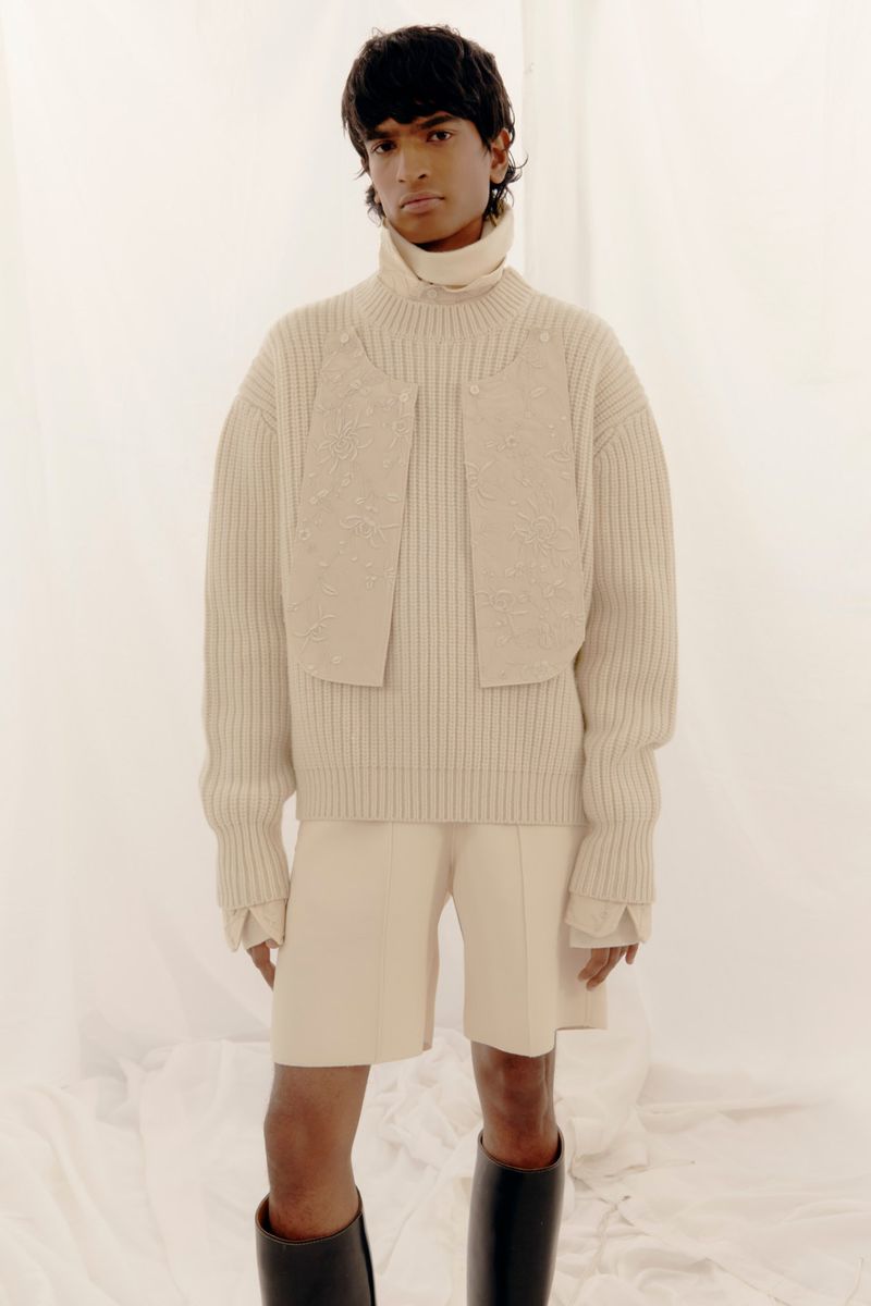 Editorial fashion shot of a person with dark hair wearing a beige ribbed sweater, shorts, and black boots against a white backdrop.