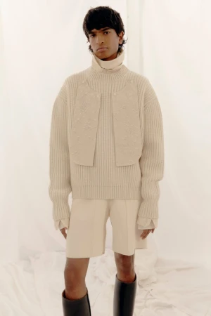 Editorial fashion shot of a person with dark hair wearing a beige ribbed sweater, shorts, and black boots against a white backdrop.