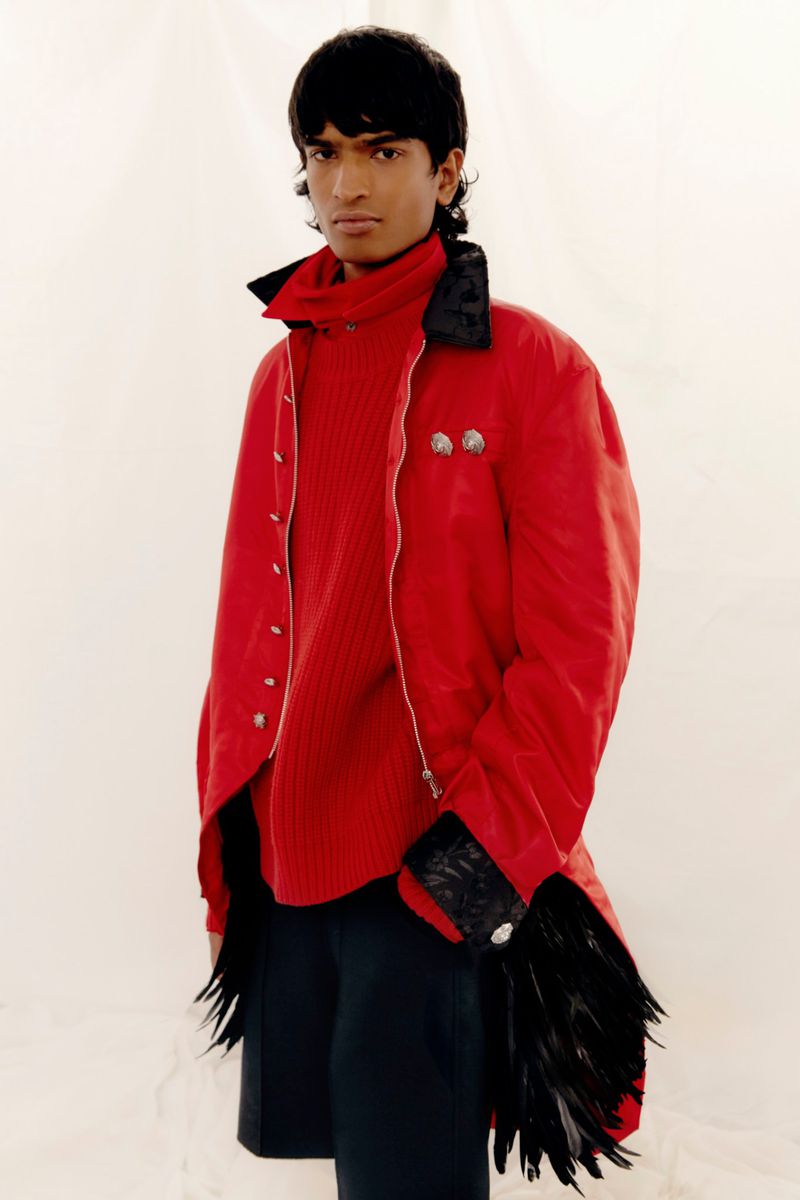 Fashionable adult male posing indoors wearing a red turtleneck sweater and red coat with feather details against a neutral backdrop.
