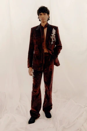 Model wearing a dark red patterned suit with embroidered brooch details against a pale backdrop