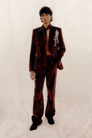 Model wearing a dark red patterned suit with embroidered brooch details against a pale backdrop