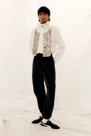 Model in white embellished shirt and black trousers standing against a textured white background.