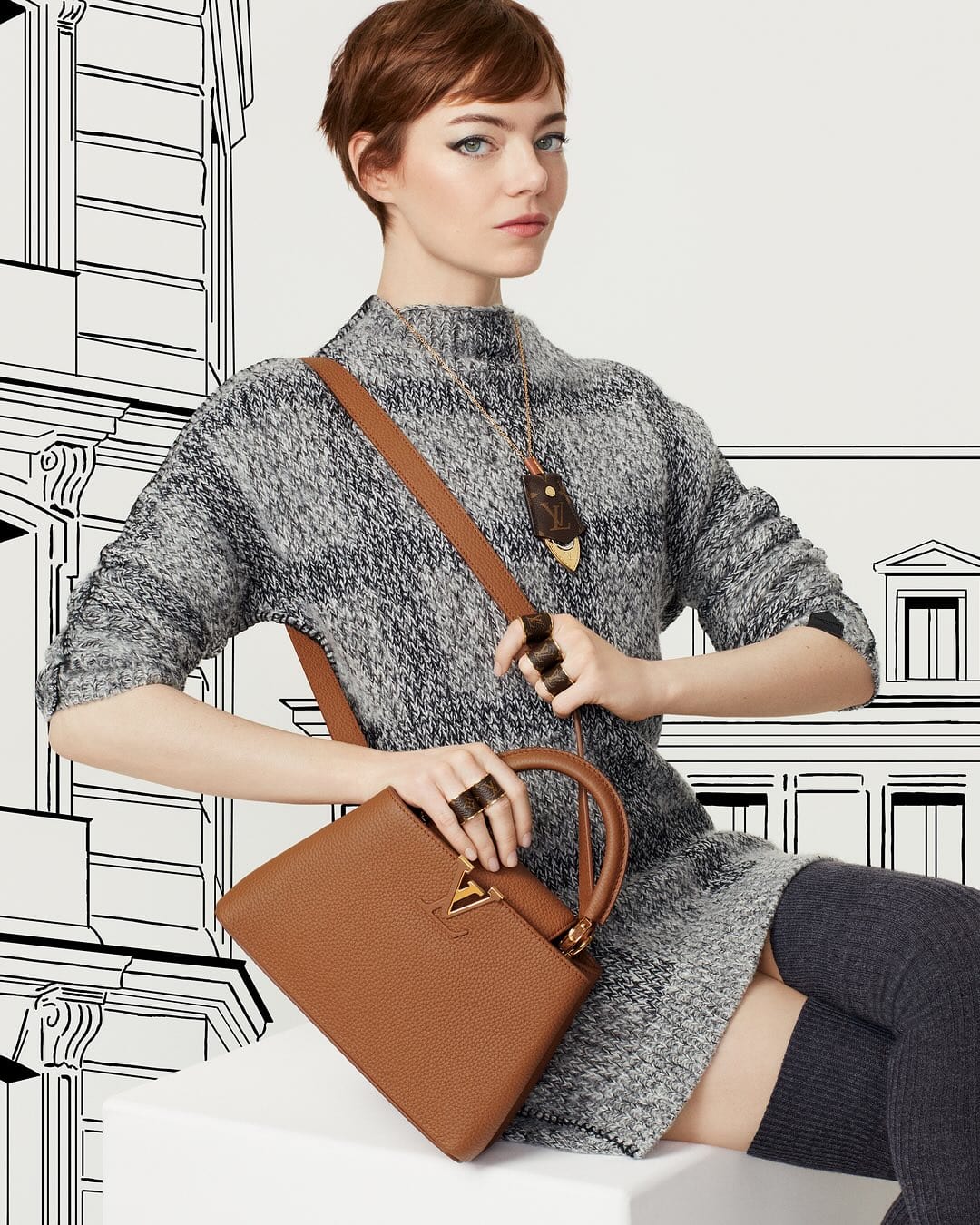 Emma Stone poses with a brown leather handbag and grey sweater dress against a white backdrop with black line art of buildings.