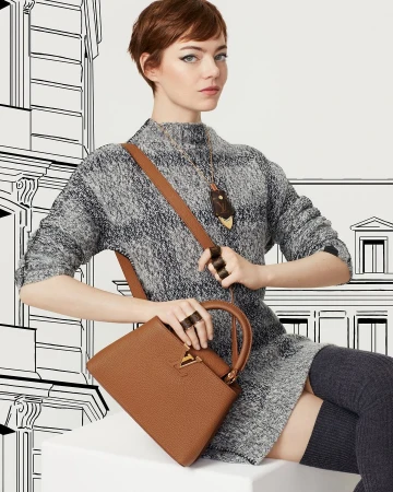 Emma Stone in a grey sweater holding a brown leather handbag against an illustrated architectural backdrop