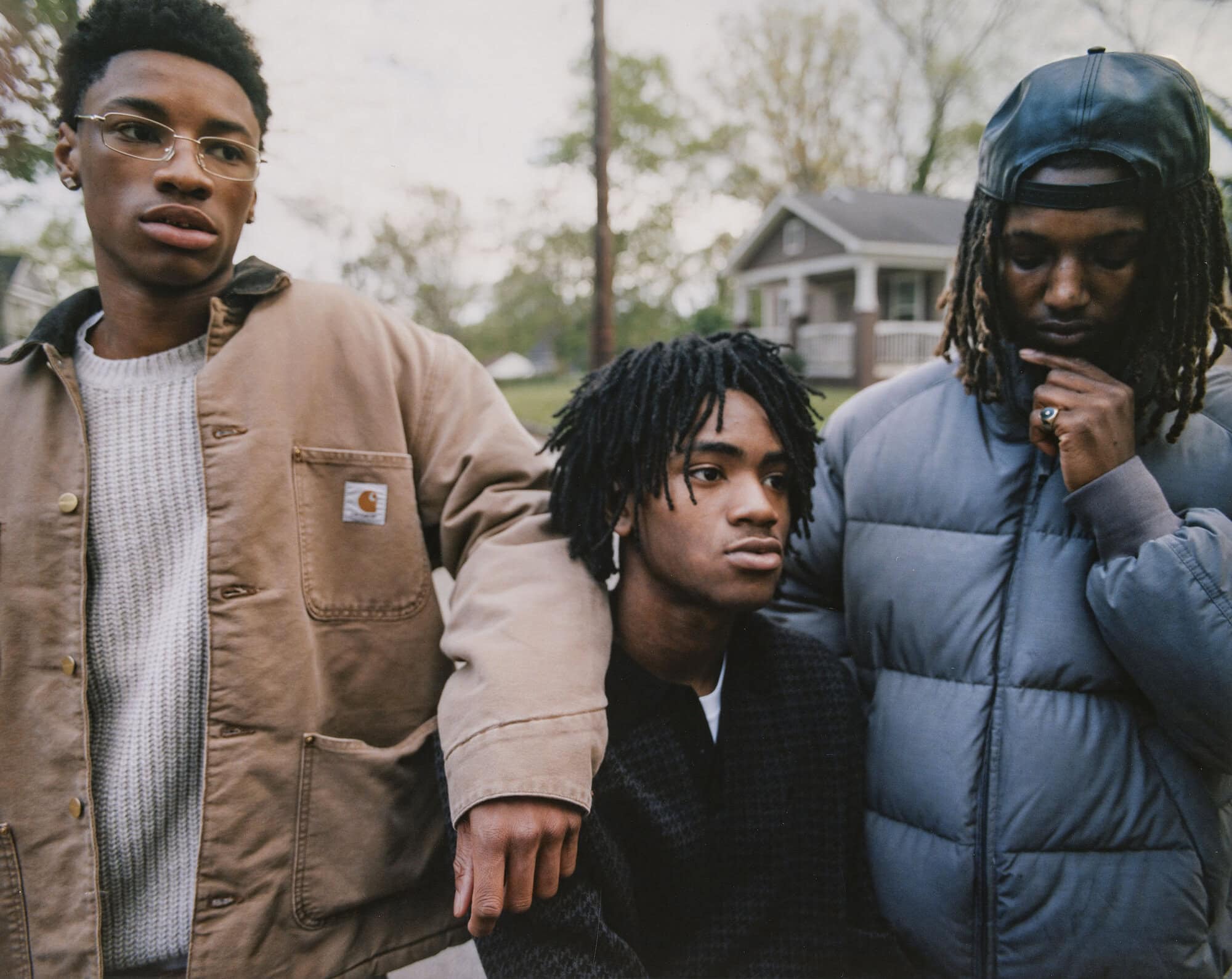 Three young African-American men pose outdoors near a house, wearing stylish jackets and sweaters, capturing a blend of modern fashion and urban style.