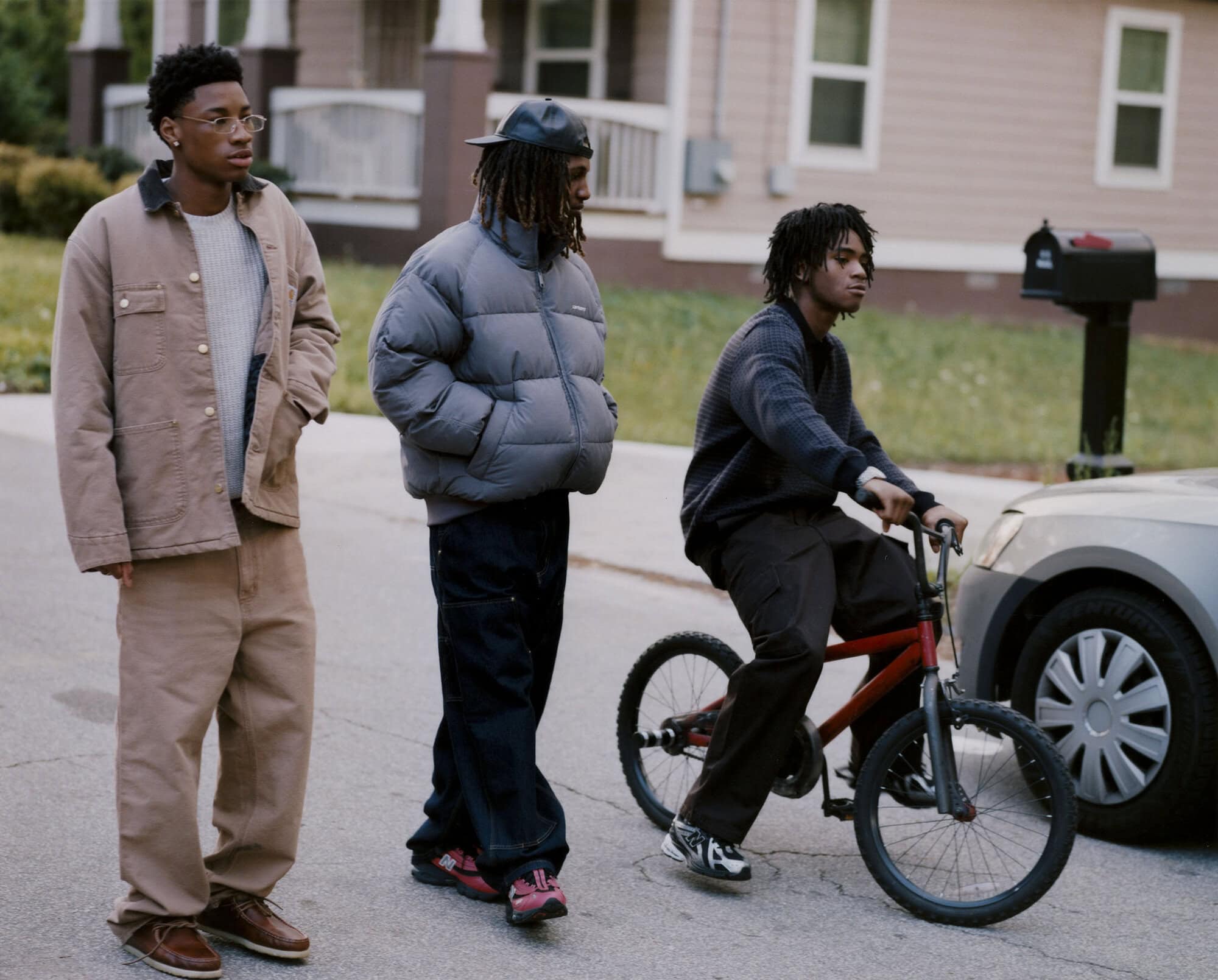 Three young adult men outdoors on a street; one rides a red bicycle, another wears a grey puffer jacket, and one wears a brown jacket.