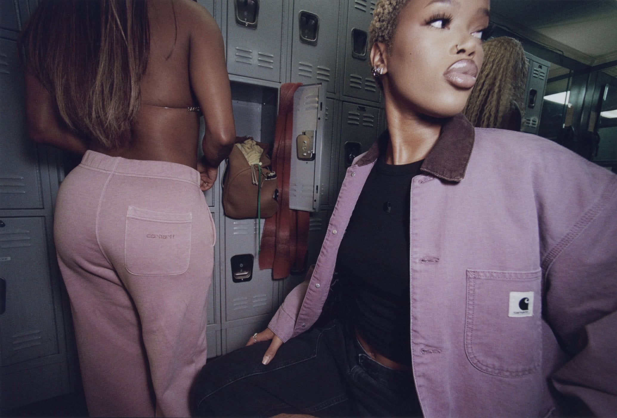 Two stylish women pose in a locker room wearing casual, coordinated outfits, showcasing fashion and lifestyle in an indoor setting.