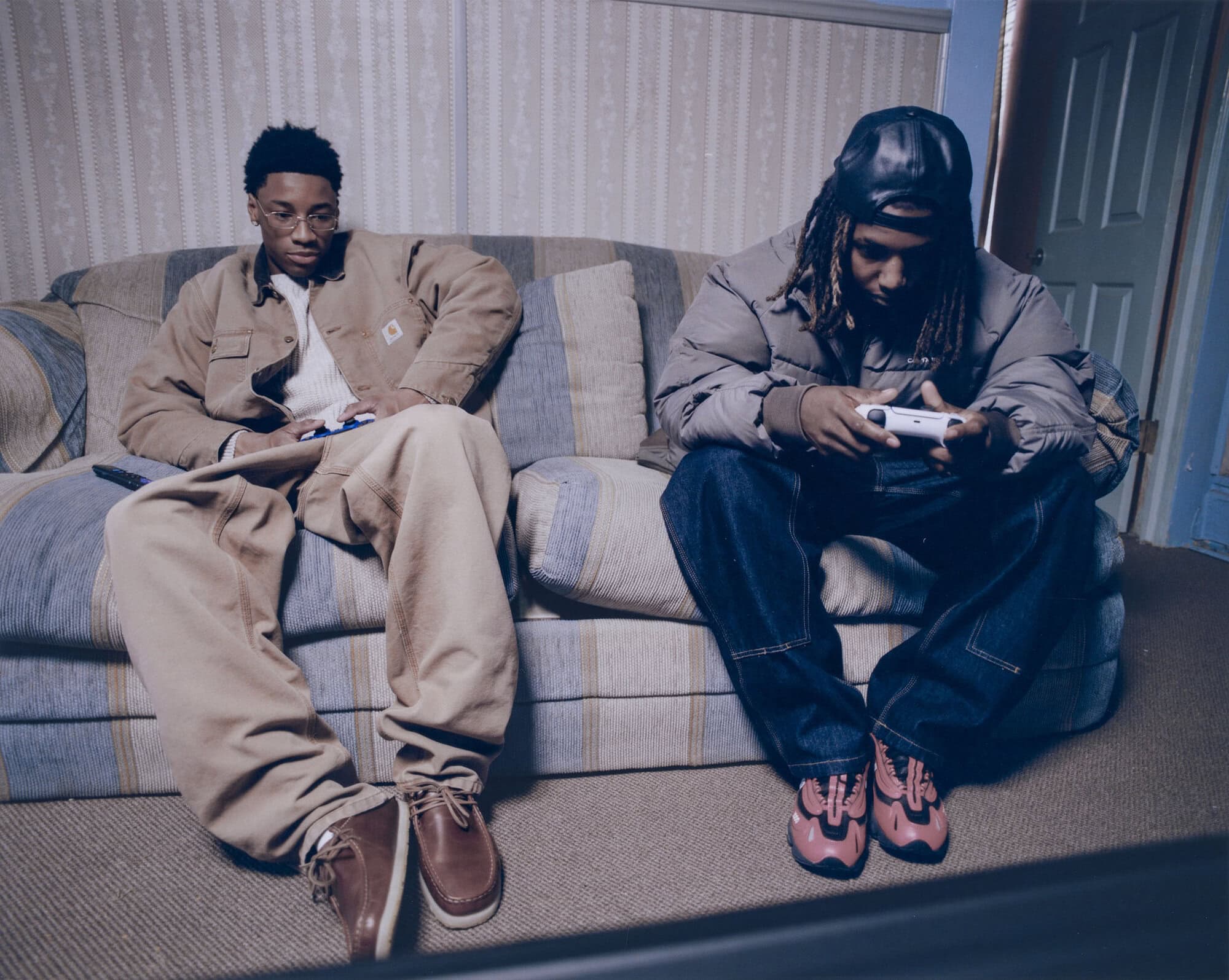 Two young men are sitting on a couch, one holding a game controller, with casual outfits and relaxed setting indoors, capturing a moment of leisure.