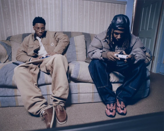 Two young men are sitting on a couch, one holding a game controller, with casual outfits and relaxed setting indoors, capturing a moment of leisure.