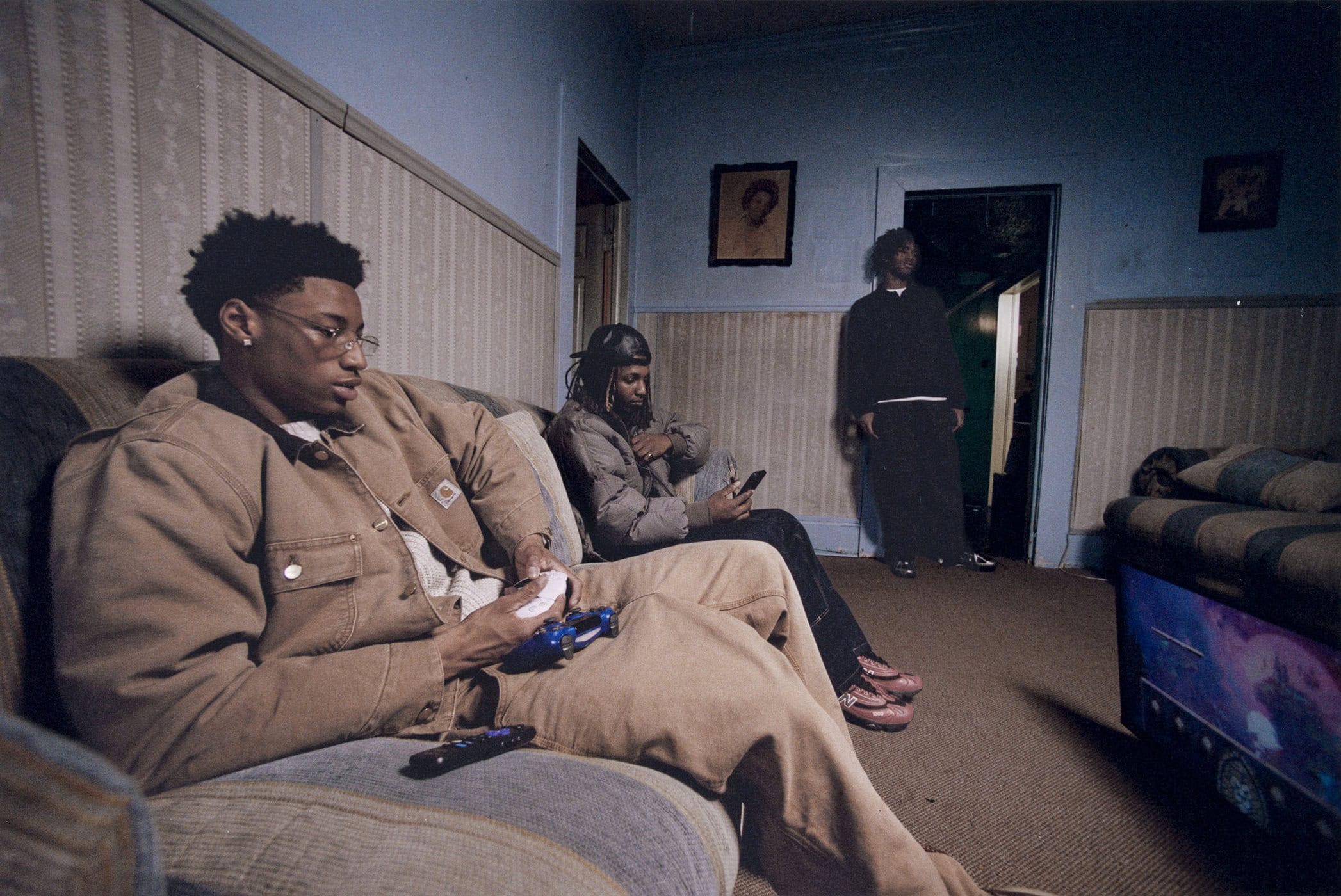 Three young men in a domestic indoor setting: two are sitting on a couch, and one is standing near a doorway in the background.