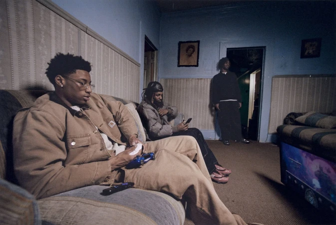 Three young men in a domestic indoor setting: two are sitting on a couch, and one is standing near a doorway in the background.