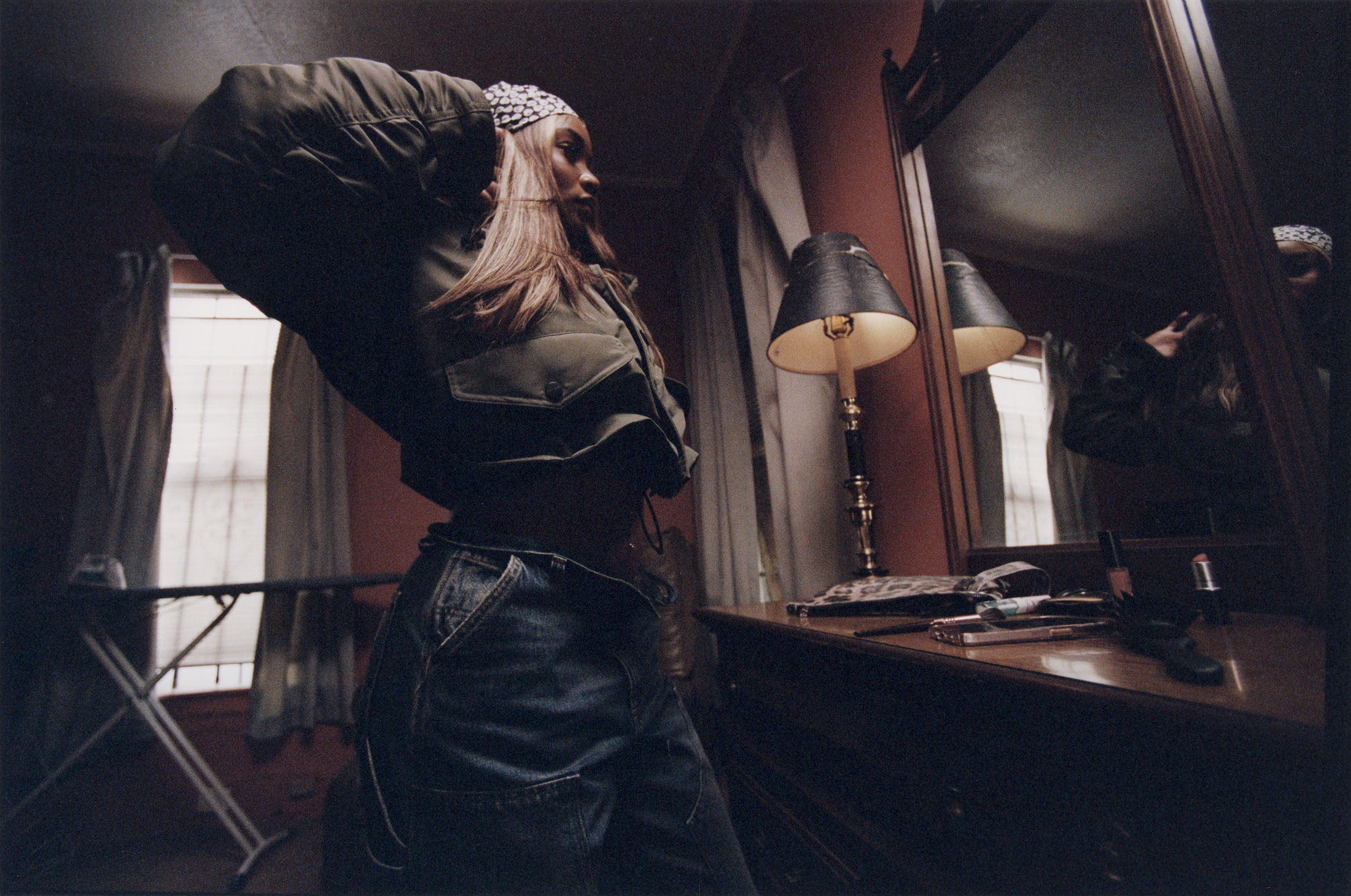A woman in a headscarf and cropped jacket adjusts her outfit in a dimly lit bedroom with a vanity, mirror, and window.
