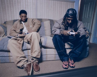 Two men seated on a patterned sofa playing video games in a living room, wearing casual streetwear and puffer jackets.
