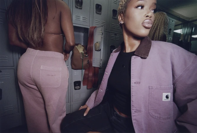 Two models inside a locker room wearing Carhartt WIP clothing, featuring a pink canvas jacket and pink sweatpants.