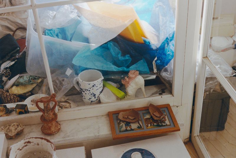 Still life of assorted objects including ceramics, framed art, and packaging inside a white-framed window, creating a cluttered, artistic display.